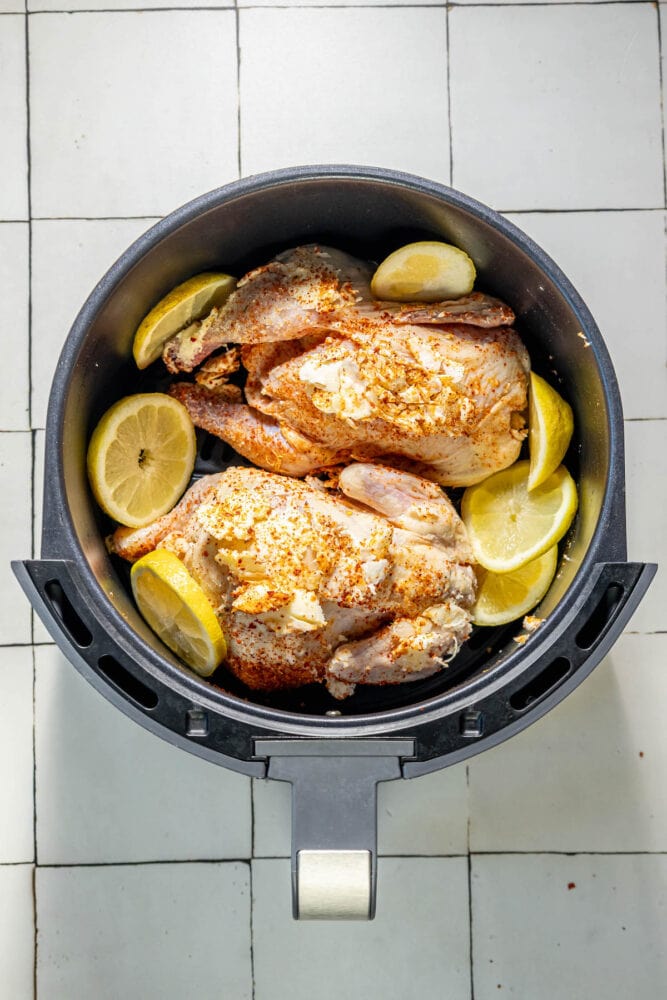raw cornish game hens in an air fryer basket with seasonings and sliced lemons