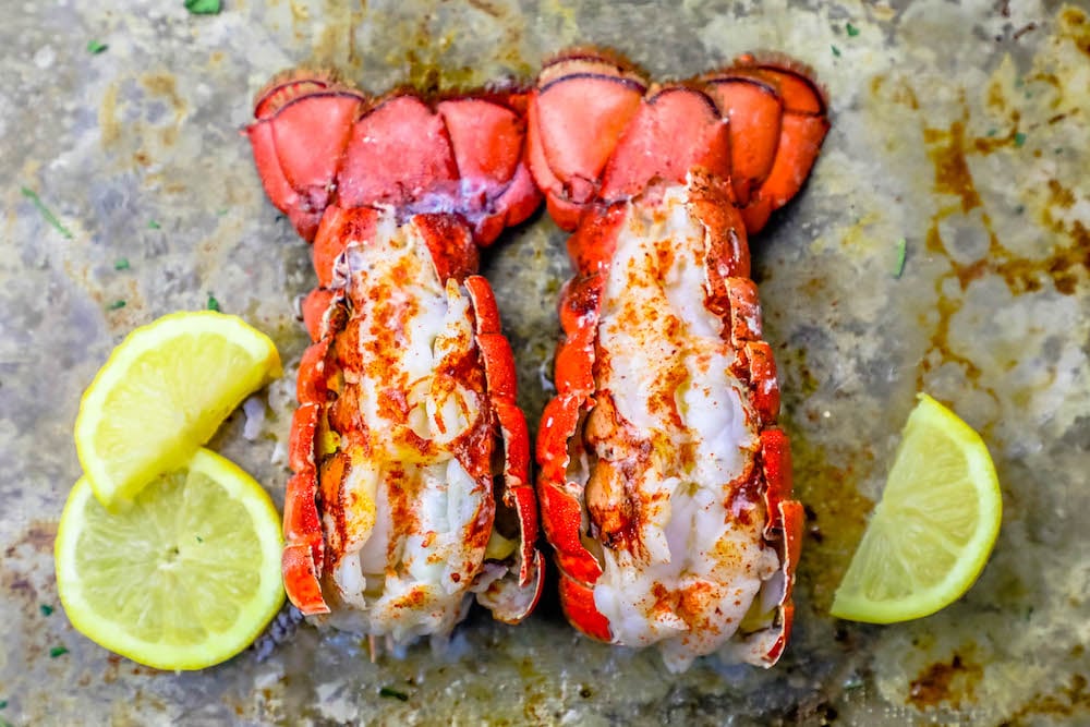two broiled lobster tails on a baking sheet with lemon slices around it